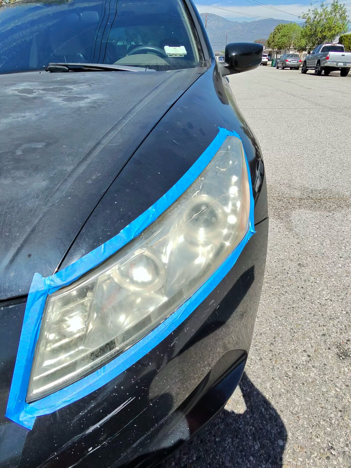 Before: yellowed, cloudy driver-side headlight on a black sedan, taped off for restoration